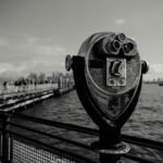 Black and white image of a coin-operated viewer on a pier overlooking the water.